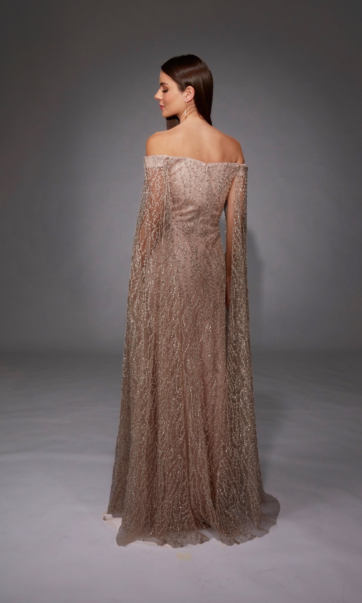A woman faces away in the Alyce 27821 long formal dress, showcasing off-the-shoulder straps, shimmering sheer fabric, and a subtle train against a plain gray background.